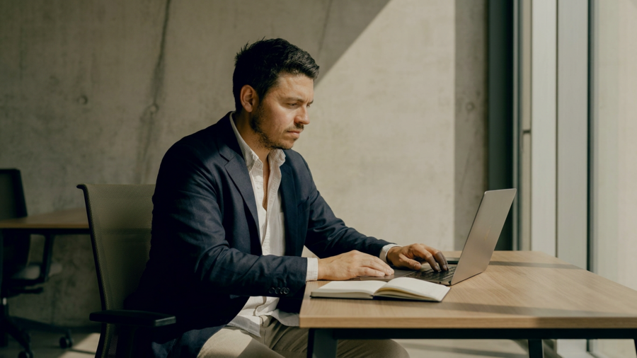 Ein Mann im Anzug sitzt vor seinem Laptop und guckt auf den Bildschirm. Hinter ihm ist eine betonfarbene Wand zusehen.