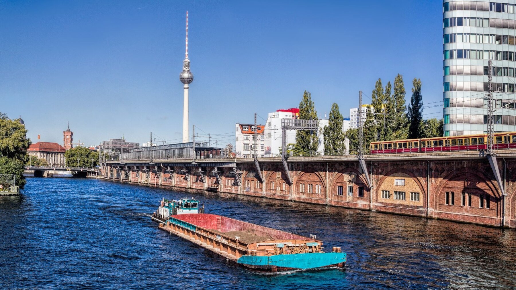 Zu sehen ist ein Fluss, auf dem ein Güterschiff fährt. Im Hintergrund sieht man einen Turm mit einer Kugel unter der Spitze. Es handelt sich um einen Spreearm in Berlin und dem Fernsehturm im Hintergrund.