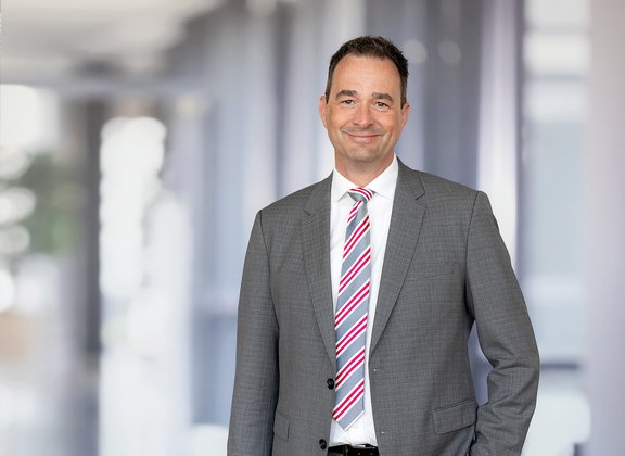 Mann in grauem Anzug, weißem Hemd und rot-weiß gestreifter Krawatte steht vor unscharfem Bürohintergrund.