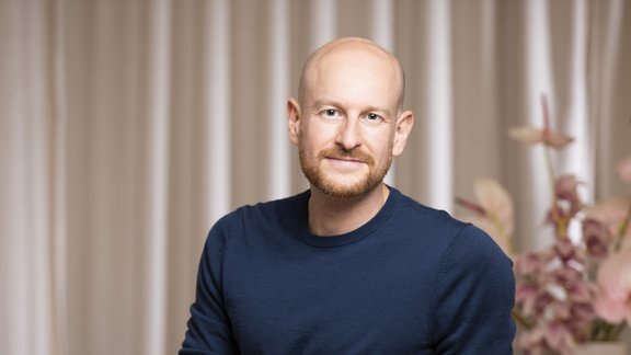 Mann mit blauem Pullover und schwarzer Hose sitzt seitlich auf einem Stuhl, im Hintergrund Vorhänge und eine Vase mit Blumen.
