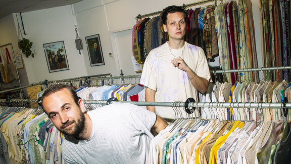 Gründer Nils Neubauer und Michael Pfeifer in einem Second Hand Geschäft mit bunter Kleidung.  