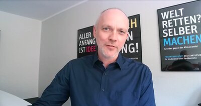 Zu sehen ist ein Mann in blauem Hemd, der in einem Zimmer sitzt. im Hintergrund hängen Poster an der Wand.