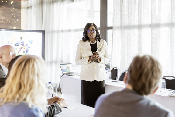 Eine Person steht bei einer Präsentation vor einem Tisch, an dem mehrere andere Personen sitzen. Hinter der stehenden Person befinden sich ein Bildschirm und ein aufgeklappter Laptop.
