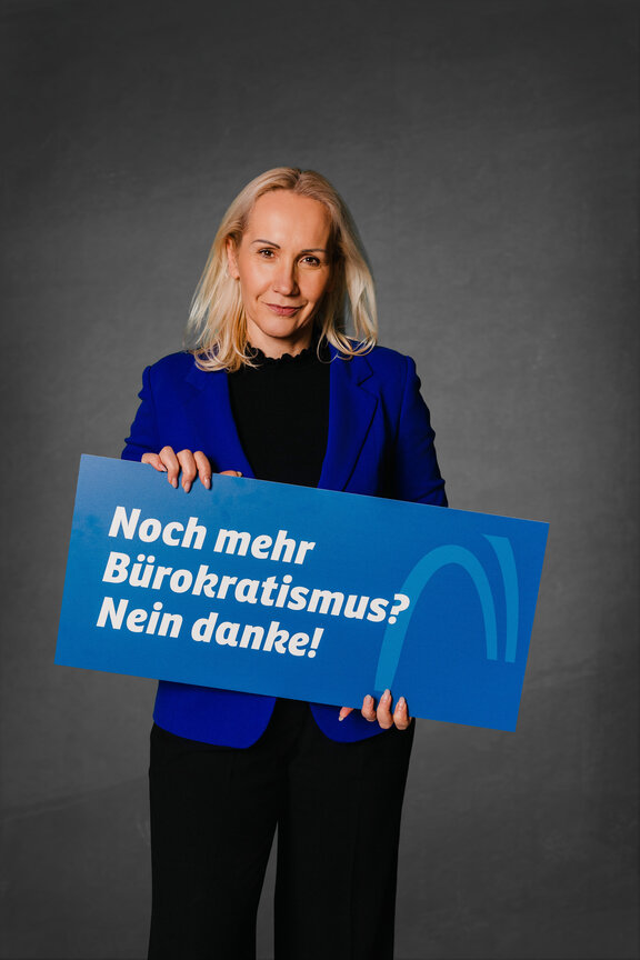 Eine Person steht vor einem neutralen grauen Hintergrund und hält ein großes blaues Schild mit der weißen Aufschrift: "Noch mehr Bürokratismus? Nein danke!"