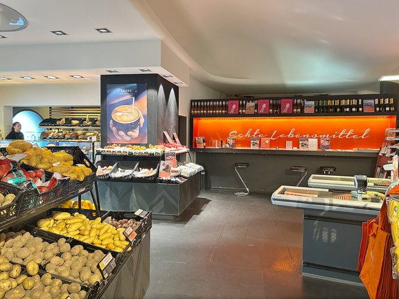 Innenansicht eines Lebensmittelgeschäfts mit Auslagen von Kartoffeln, Brot und anderen Lebensmitteln, beleuchtetes rotes Schild mit Schriftzug 'Echte Lebensmittel'