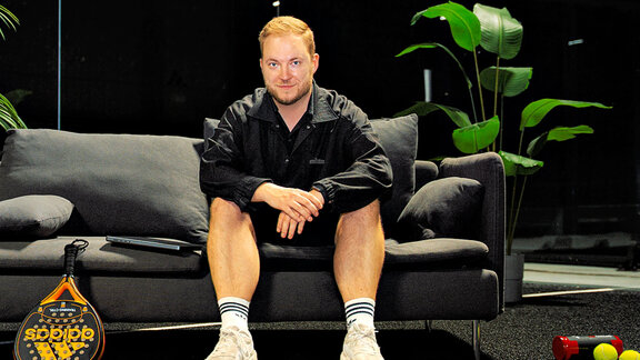 Ein Mann sitzt auf einem Sofa und lächelt in die Kamera. Es ist Bastian Krautwald von The Padel Company