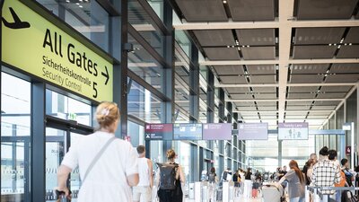 Zu sehen sind diverse Menschen, die in einem großen Gebäude mit Koffern einen Gang entlanglaufen. Es ist eine Szene aus dem Flughafen BER.