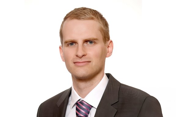 Mann mit kurzem blondem Haar trägt dunklen Anzug, weißes Hemd und gestreifte Krawatte in Blau und Pink.