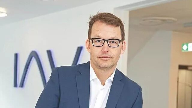 Person in einem dunkelblauen Anzug steht in einem modernen Bürobereich mit dem IAV‑Logo im Hintergrund
