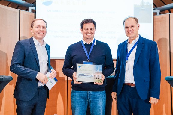 Drei Männer in Business-Kleidung, der mittlere hält eine Auszeichnung mit blauem Band, alle lächeln in einem Konferenzraum