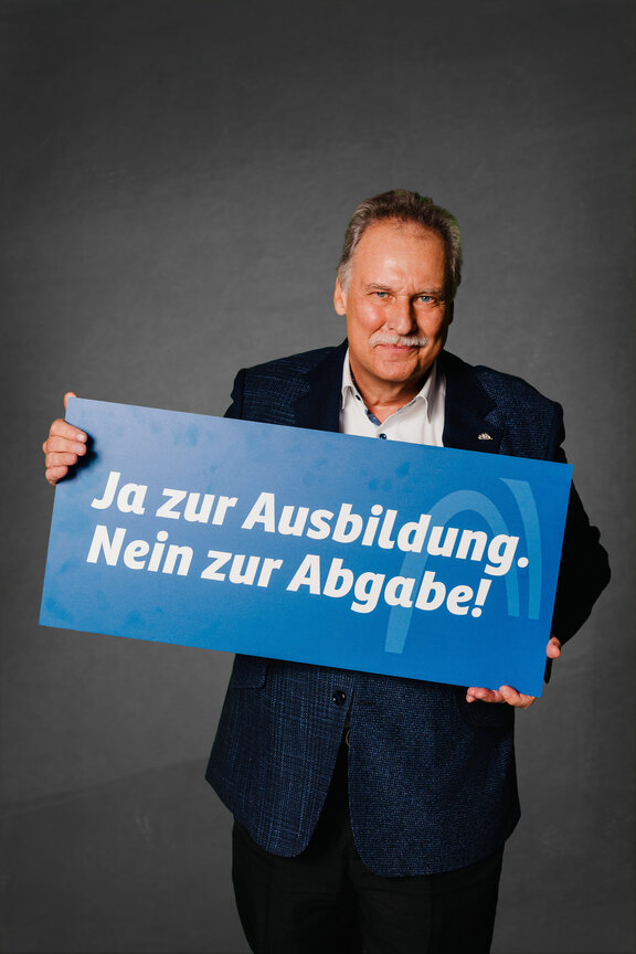 Eine Person steht vor einem neutralen grauen Hintergrund und hält ein großes blaues Schild mit der weißen Aufschrift: "Ja zur Ausbildung. Nein zur Abgabe!"