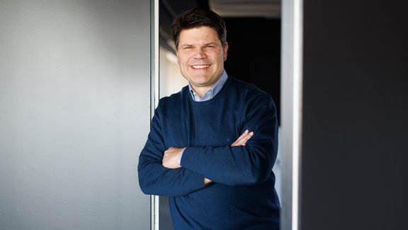 Mann mit dunkelblauem Pullover und hellblauem Hemd steht mit verschränkten Armen in einem Türrahmen.