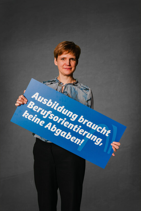 Eine Person steht vor einem neutralen grauen Hintergrund und hält ein großes blaues Schild mit der weißen Aufschrift: "Ausbildung braucht Berufsorientierung, keine Abgabe!"
