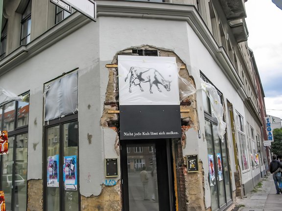 Ecke eines Gebäudes mit beschädigter Fassade, darüber ein Schild mit einer Kuhzeichnung und dem Text 'Nicht jede Kuh lässt sich melken'