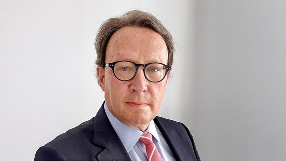 Mann mit mittellangem, graubraunem Haar trägt dunkelblauen Anzug, hellblaues Hemd und rot-weiß gestreifte Krawatte, steht vor weißer Wand