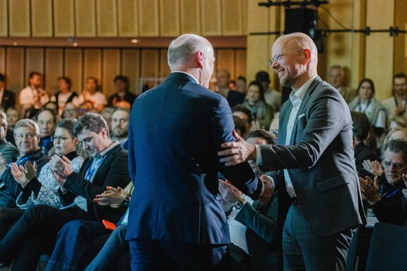 Zwei Männer reichen sich auf einer Bühne vor Publikum freudig die Hand.