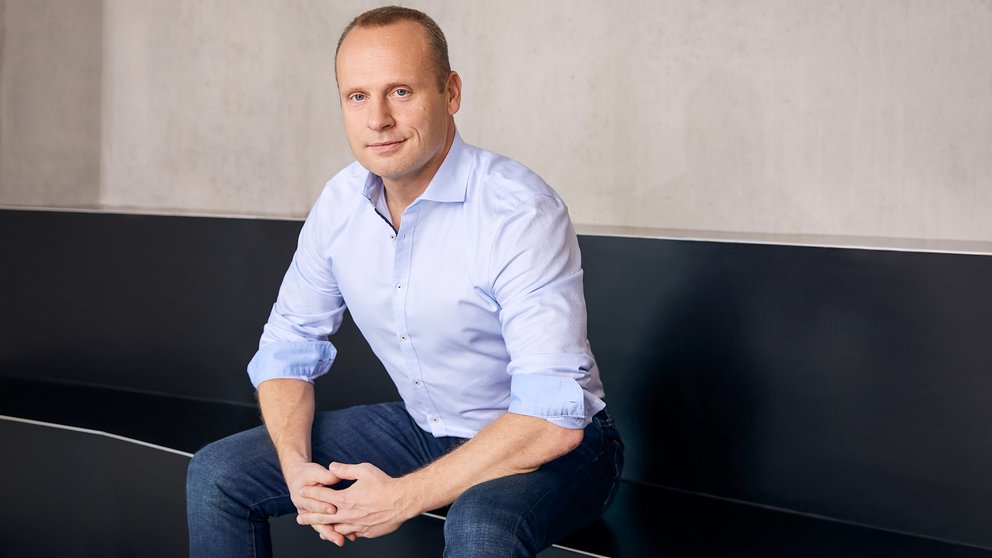 Mann in einem hellblauen Hemd sitzt mit zusammengelegten Händen auf einem schwarzen Sofa. 
