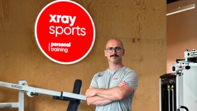 Ein Mann mit einer Brille steht mit verschrenkten Armen vor einem roten Leuchtschild und Sportgeräten