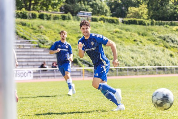 Zwei Fußballspieler in blauen Trikots laufen auf einem Spielfeld, einer im Vordergrund in Bewegung, der andere im Hintergrund