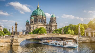 Zu sehen ist ein Fluss, über den eine Brücke führt. unter der Brücke hindurch fährt ein Boot. im Hintergrund ein imposantes Gebäude, der Berliner Dom.
