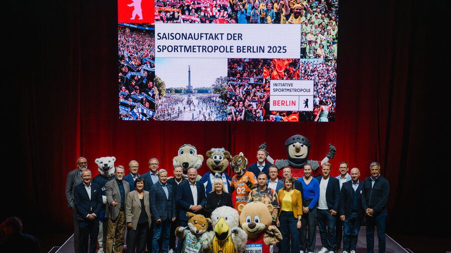 Man schaut auf enie Bühne, auf der viele Menschen stehen und für ein gemeinsames Foto posieren. Hinter der Bühne ist ein roter Vorhand und davor eine große Leinwand. Es handelt sich um die Presseveranstaltung aus dem Jahre 2025 zum Thema Sportmetropole Berlin.