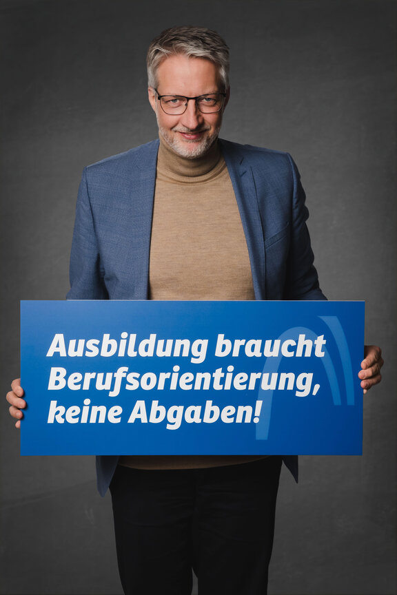 Eine Person steht vor einem neutralen grauen Hintergrund und hält ein großes blaues Schild mit der weißen Aufschrift: "Die Strafabgabe schwächt den Wirtschaftsstandort!"
