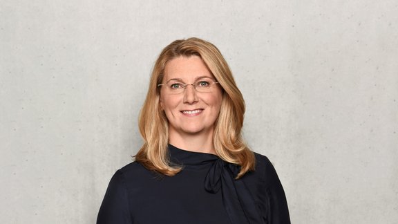 Frau mit schulterlangem, blondem Haar trägt eine schwarze Bluse mit Schleife am Kragen vor grauer Wand.