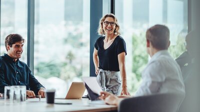 Zu sehen ist ein Büroraum. Drei Personen sitzen bzw. stehen um den Tisch und haben eine fröhliche Mimik.