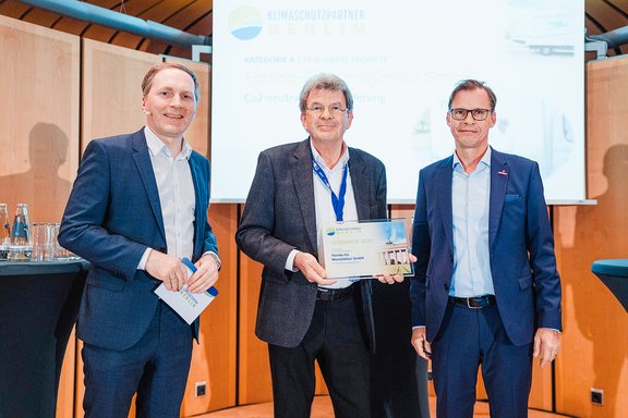Drei Männer in Anzügen bei einer Preisverleihung, der mittlere hält eine Urkunde, Hintergrund mit Präsentationsfolie