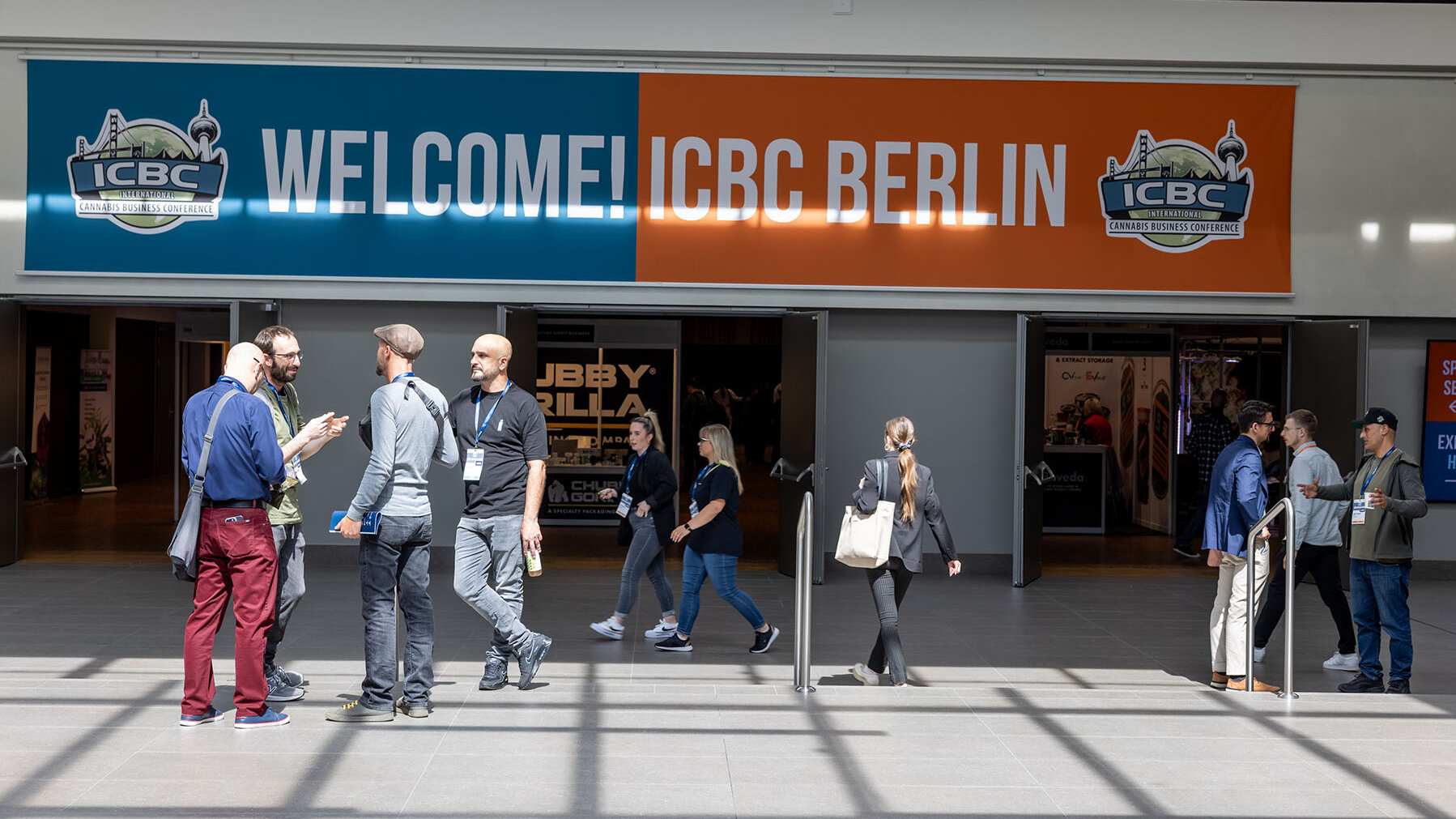 Zu sehen sind mehrere Personen. die vor dem Eingang eines Messe-Gebäudes stehen, hinein- oder hinausgehen oder sich unterhalten. Über dem Eingang hängt ein Banner mit der Aufschrift: Welcome ICBC Berlin. Es handelt sich um eine Szene auf der Cannabismesse ICBC.