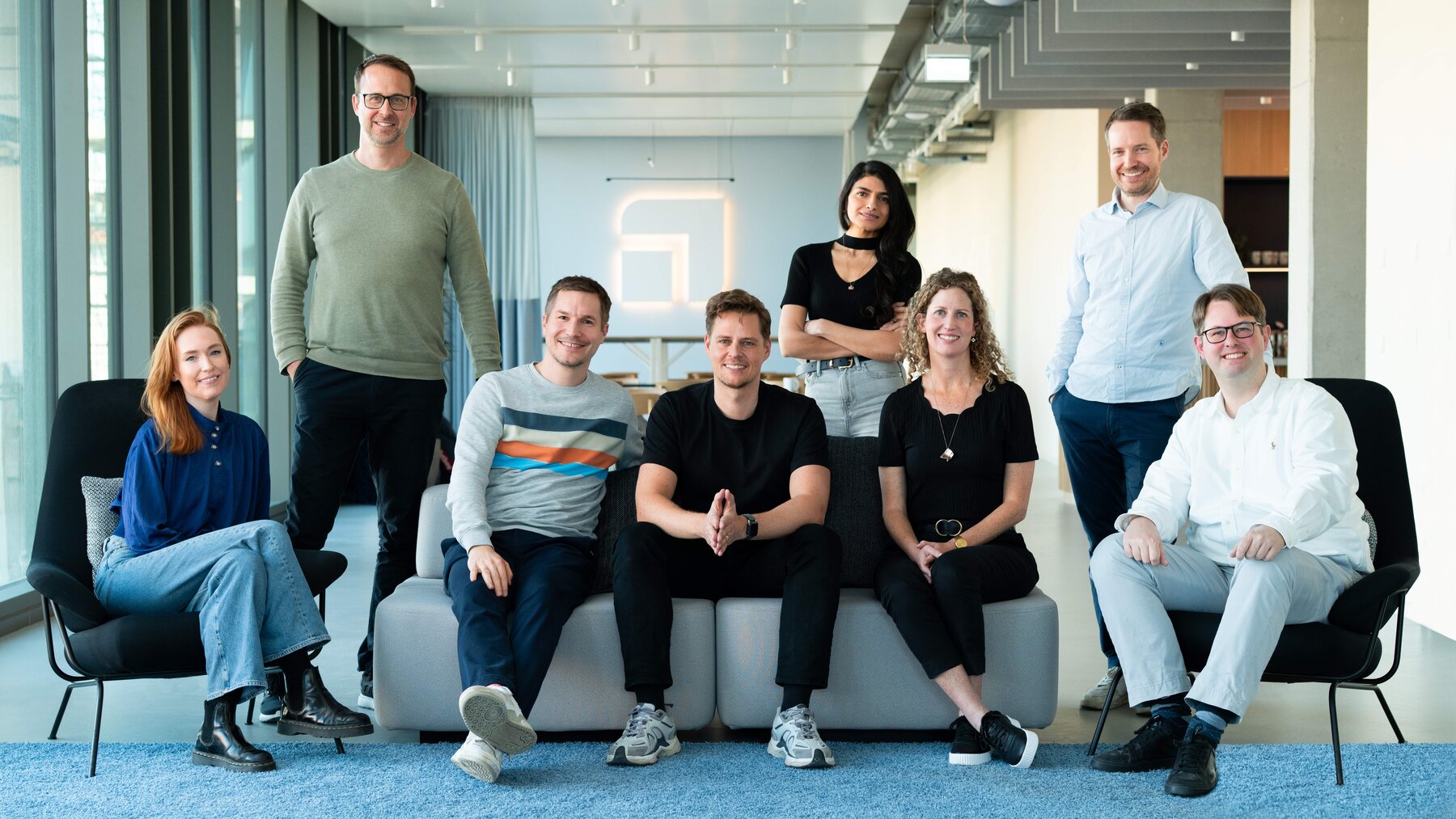 8 Menschen stehen verteilt um ein Sofa herum. 3 der Personen sitzen auf dem Sofa 