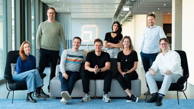 8 Menschen stehen verteilt um ein Sofa herum. 3 der Personen sitzen auf dem Sofa 