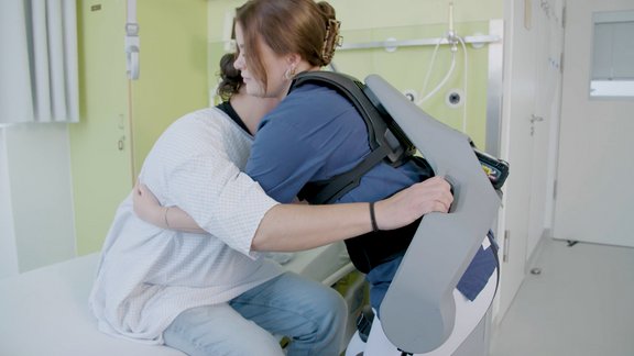 Eine Frau mit einem Exoskelett versucht eine andere Personen beim Aufstehen von einem Bett zu helfen.