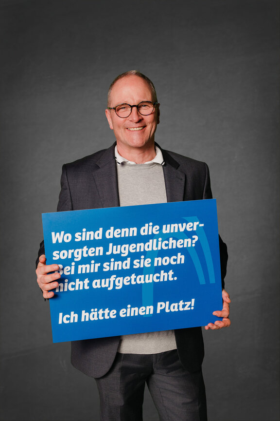 Eine Person steht vor einem neutralen grauen Hintergrund und hält ein großes blaues Schild mit der weißen Aufschrift: "Ich finde keine Azubis und muss dann ein Abgabe zahlen. Das ist doppelt bestraft!"