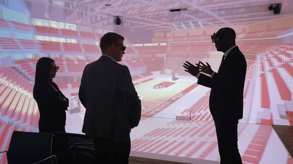 Drei Personen in Anzügen tragen 3D-Brillen und sehen eine Plansimulation eines Stadions