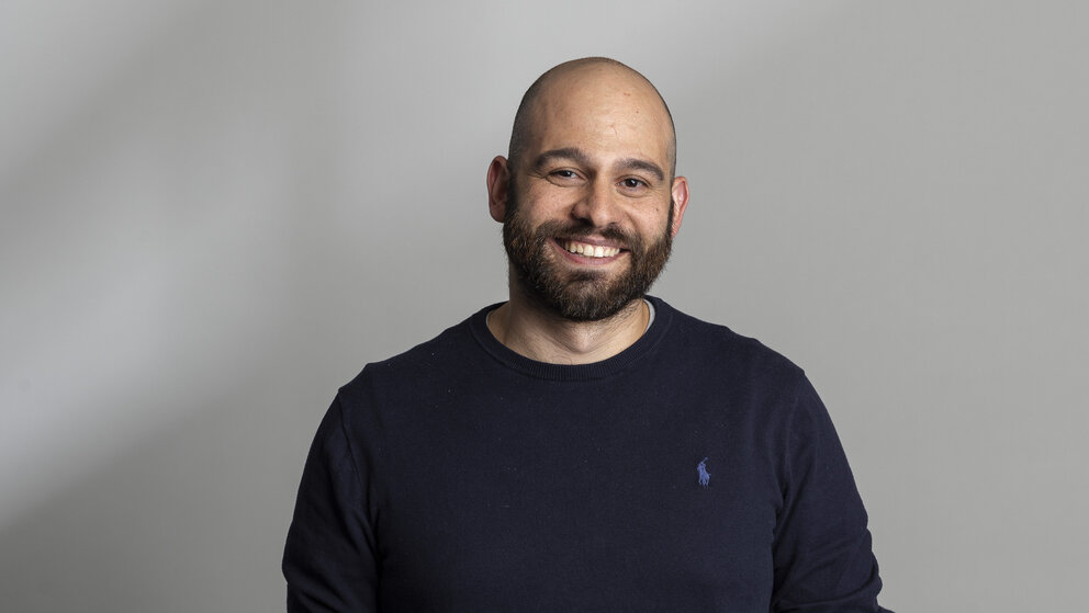 Baha Jamous, Director of People and Corporate Affairs bei Solaris, steht in dunklem Pullover und beiger Hose lachend vor einem hellen Hintergrund