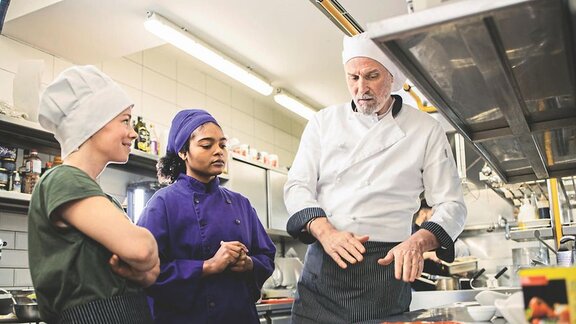 Foto von zwei jungen Menschen in einer Küche, welche von einem Koch angeleitet werden.