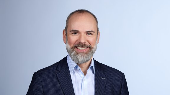 Mann mit kurzem Bart, trägt einen dunkelblauen Anzug und ein hellblaues Hemd, steht aufrecht vor einem hellen Hintergrund.