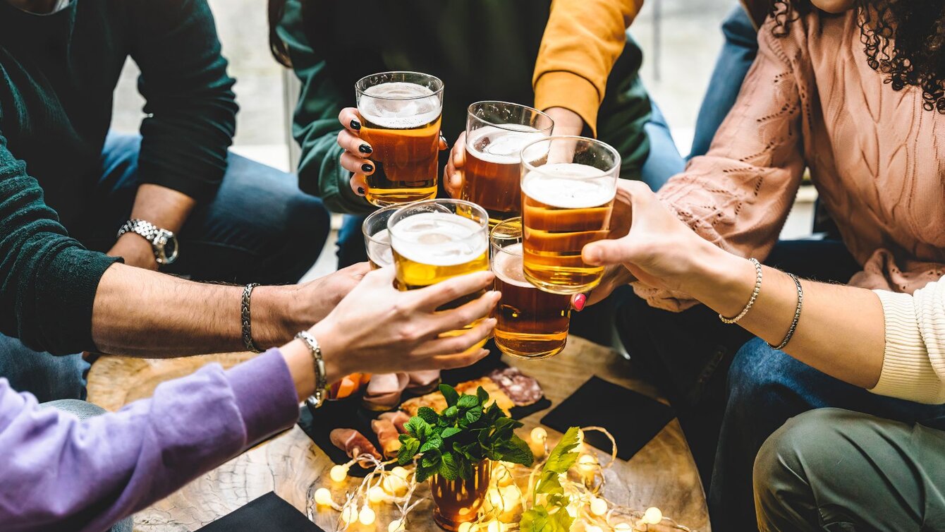 Zu sehen ist eine Freundesgruppe, die an einem Tisch sitzt und mit Biergläsern anstößt.