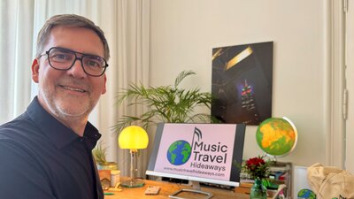 Ein Mann sitzt vor seinem Schreibtisch. Neben seinem Bildschirm ist eine gelbe Lampe und ein Globus. Er guckt in die Kamera und lächelt 