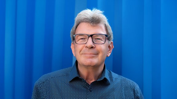 Mann mit grauen Haaren, zurückgebunden, trägt ein dunkelblaues Hemd mit weißem Punktmuster vor blauem Hintergrund.