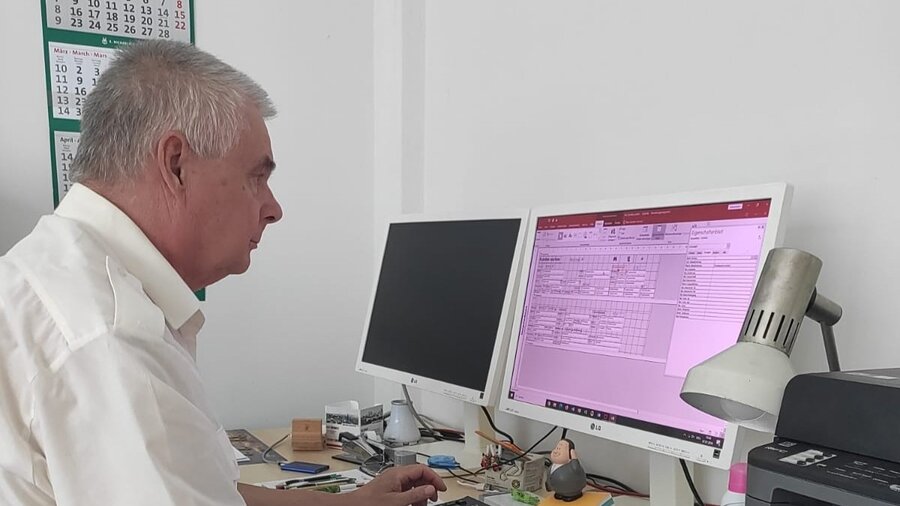 Auf dem Bild sieht man einen älteren Mann, der ein weißes Hemd trägt. Er sitzt vor einem Computer.
