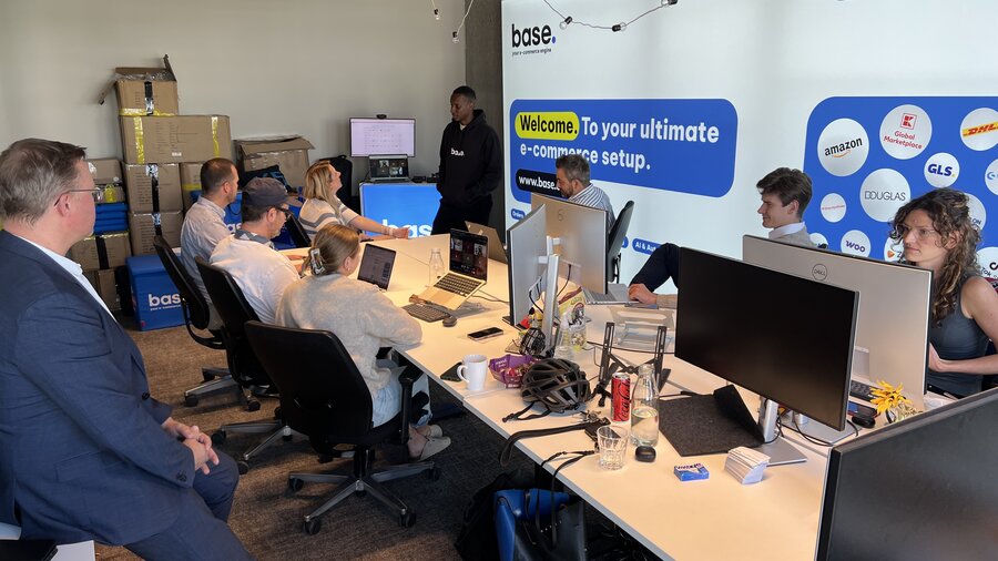 Auf dem Bild sind einige Menschen zu sehen, die gemeinsam um mehrere Schreibtische herum sitzen und am Computer arbeiten. 