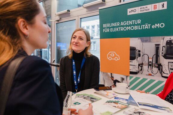 Eine Frau sitzt an einem Stand mit Werbematerialien, eine Interessierte sieht sich diese an.