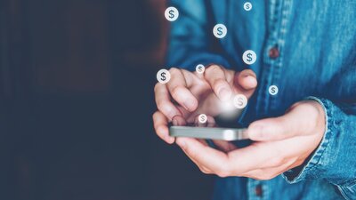 Zu sehen ist ein Handy, welches in der Hand gehalten wird, über dem Bildschirm schweben Dollar-Zeichen.