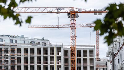 Zu sehen ist ein sich im Bau befindendes Mehrfamilien-Wohnhaus. Der Rohbau steht fast, es steht jeweils ein großer gelber Baukran vor und hinter dem Gebäude.