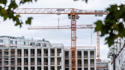 Zu sehen ist ein sich im Bau befindendes Mehrfamilien-Wohnhaus. Der Rohbau steht fast, es steht jeweils ein großer gelber Baukran vor und hinter dem Gebäude.