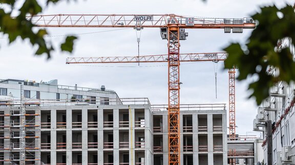 Zu sehen ist ein sich im Bau befindendes Mehrfamilien-Wohnhaus. Der Rohbau steht fast, es steht jeweils ein großer gelber Baukran vor und hinter dem Gebäude.
