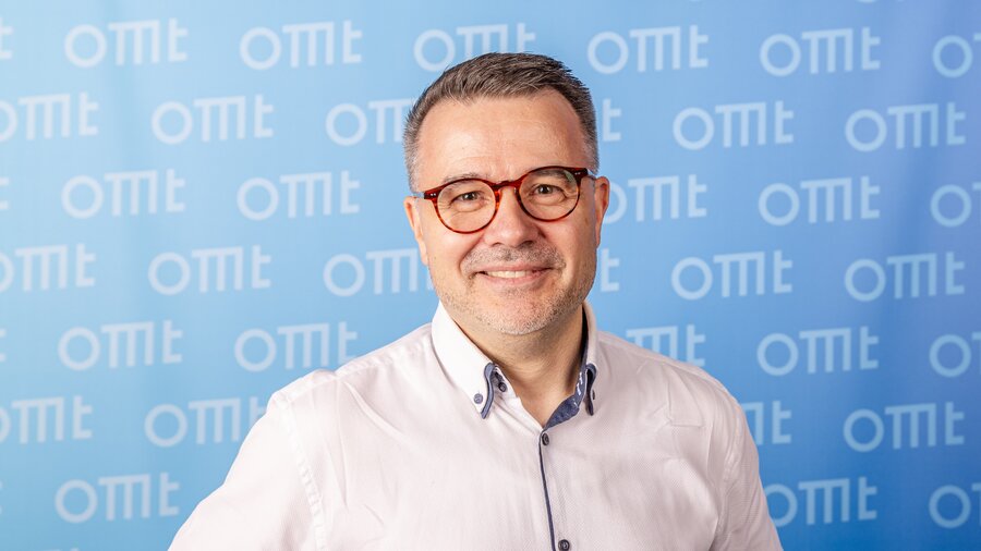 Auf dem Bild ist ein Mann mit Brille zu sehen, der vor einem blauen Hintergrund steht.