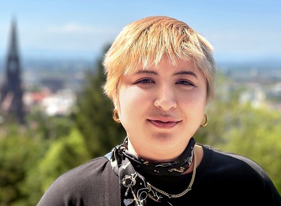 Person mit kurzen blonden Haaren, trägt schwarzen Pullover, goldene Kette und Ohrringe, im Hintergrund Stadt Freiburg mit Kirchturm